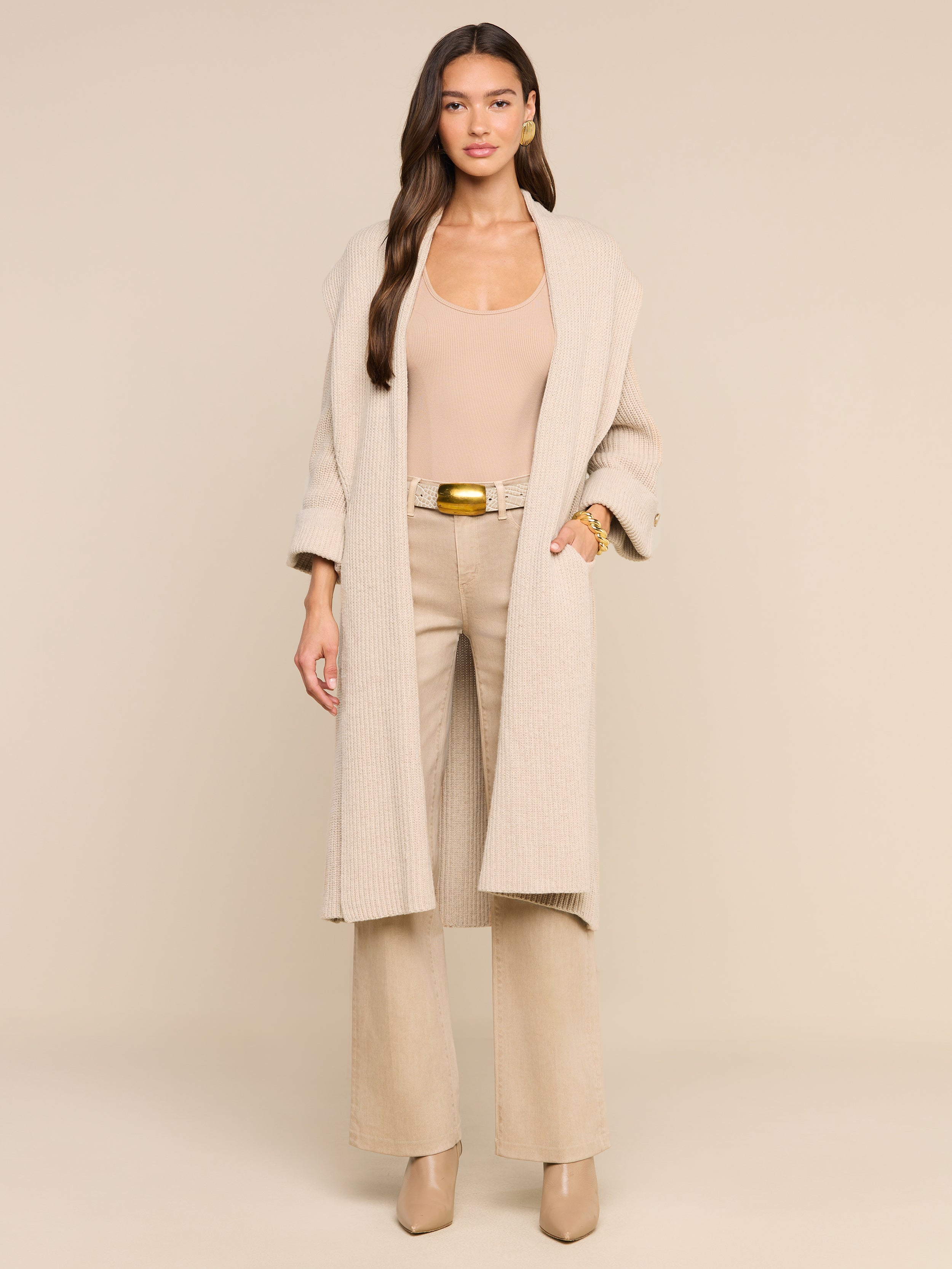 A woman with long brown hair poses against a plain white backdrop, wearing neutral accessories and heeled shoes. She layers the Orielle Cardigan over a beige top and matching pants for a coordinated studio look.