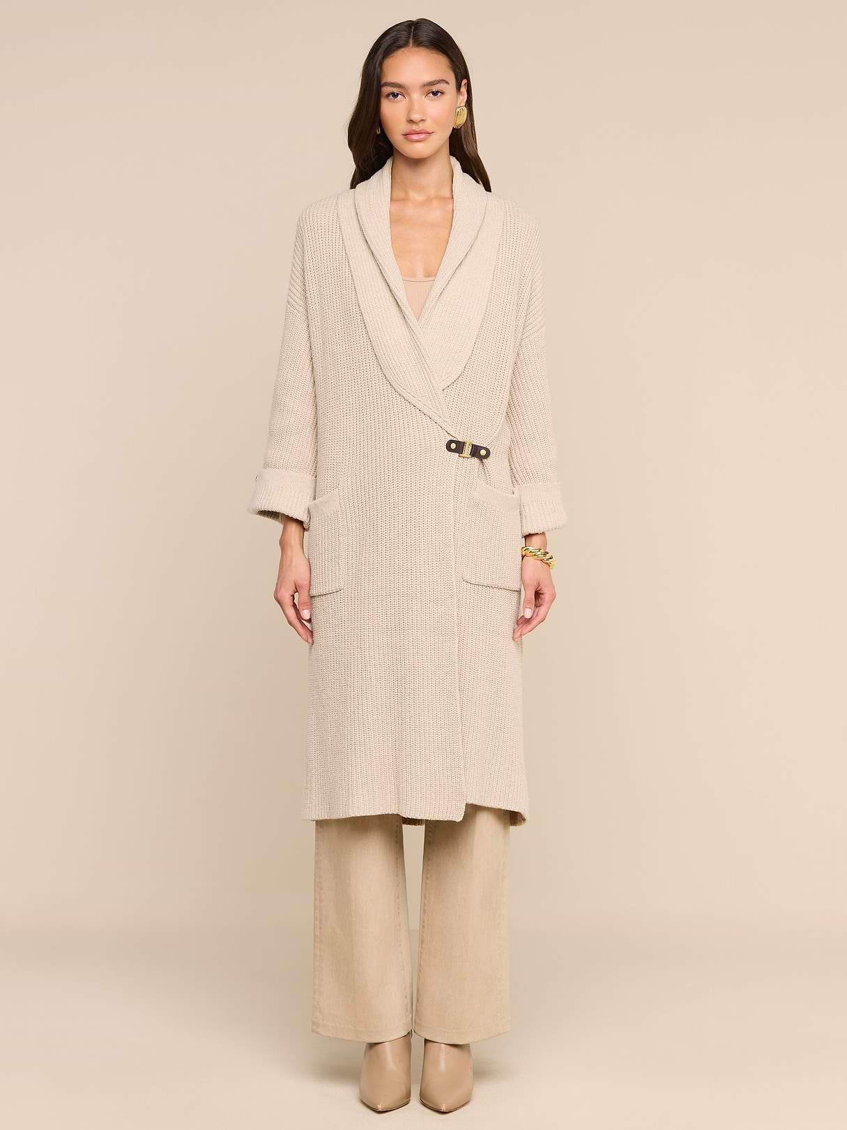 A woman stands facing forward against a plain light background, elegantly styled for fall in a beige long coat with a single clasp and wide lapels, wide-leg pants, heeled shoes, and the Orielle Cardigan layered underneath.