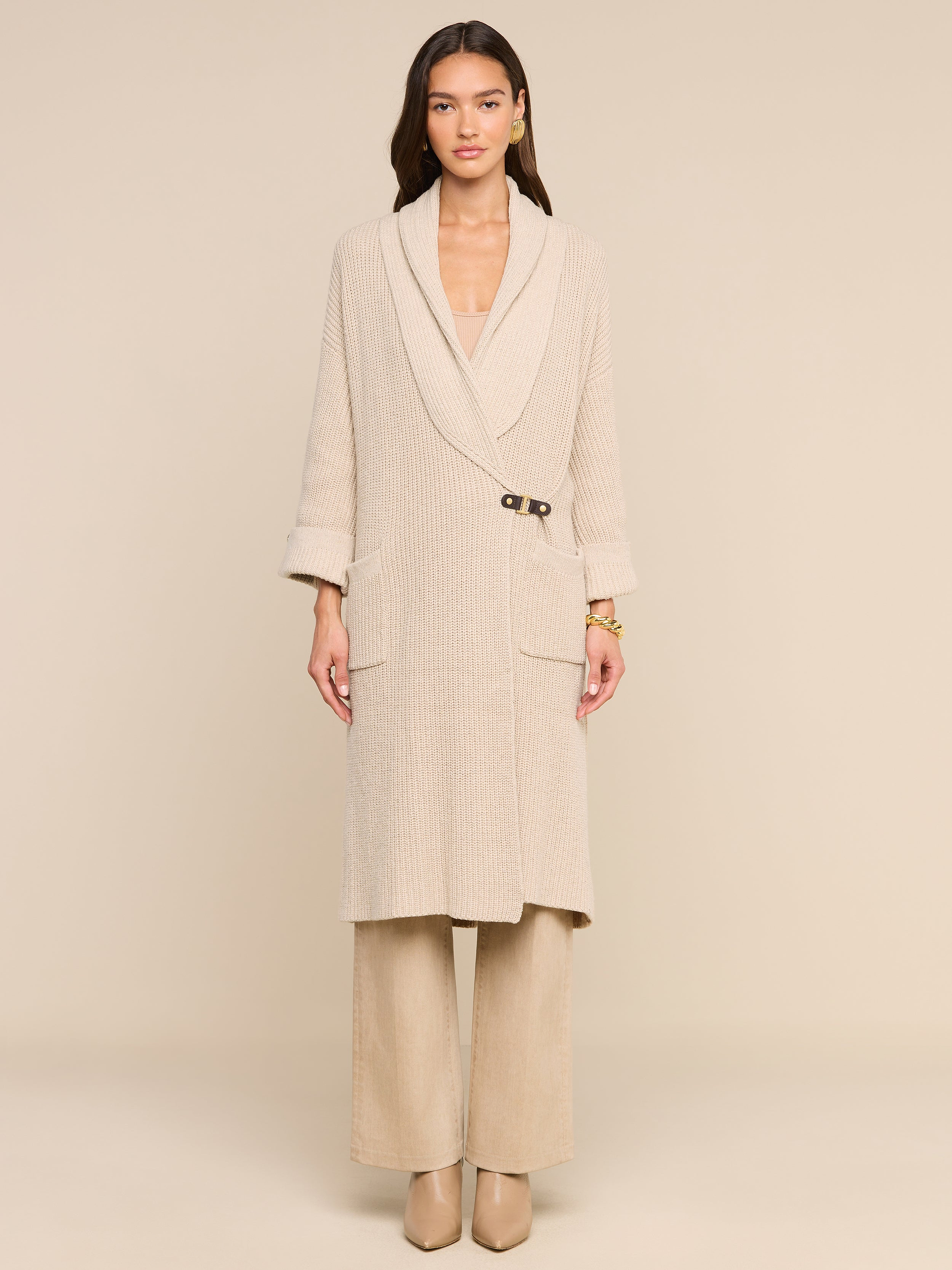 A woman stands facing forward against a plain light background, elegantly styled for fall in a beige long coat with a single clasp and wide lapels, wide-leg pants, heeled shoes, and the Orielle Cardigan layered underneath.