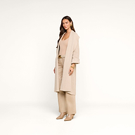 A woman with long brown hair stands against a plain white background, exuding relaxed confidence in her beige long coat, wide-leg pants, matching top, and heeled shoes. She effortlessly completes her look by layering with the Orielle Cardigan for added warmth and style.