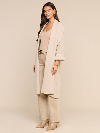 A woman stands against a beige background wearing beige wide-leg pants with a matching belt, a tan top, heeled boots, gold jewelry, and the Orielle Cardigan styled as a long cream fall sweater.