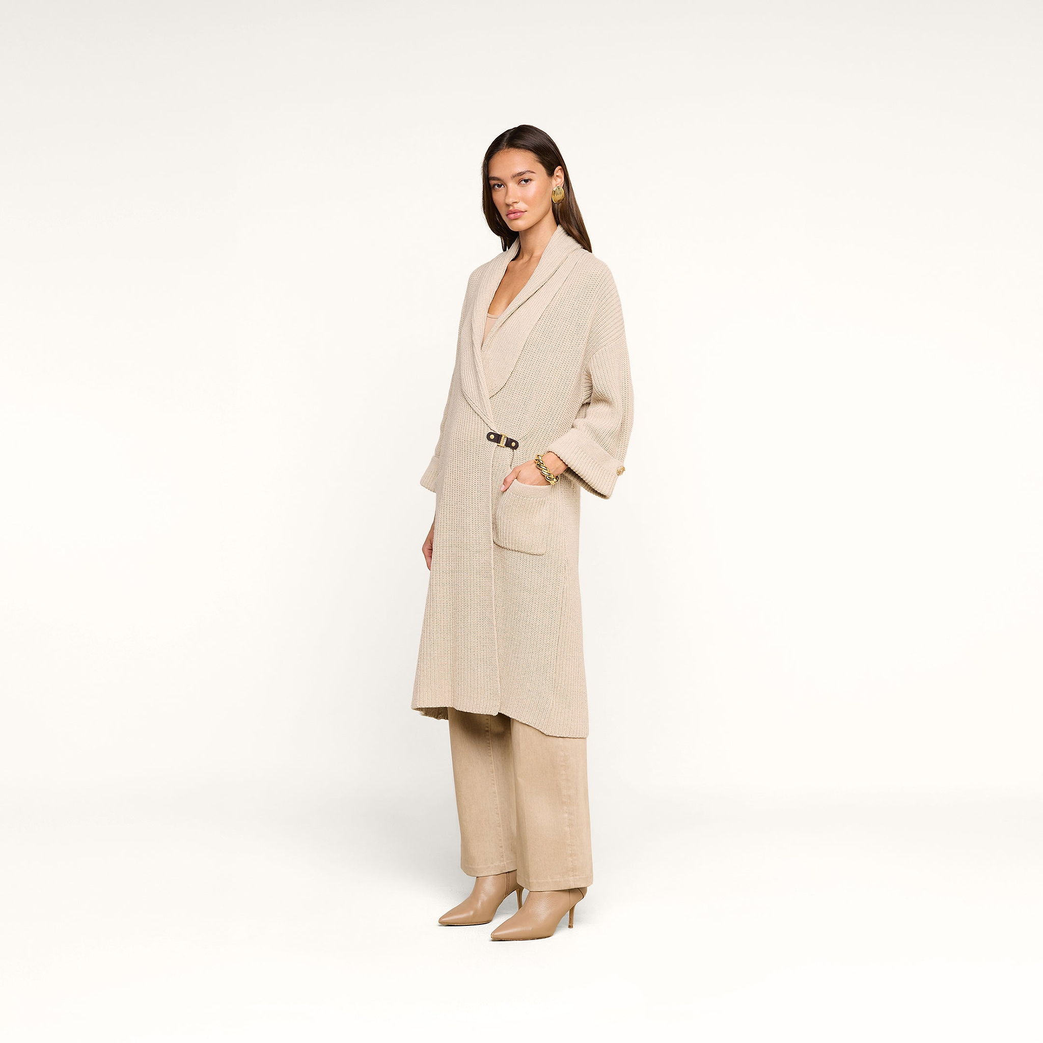 A woman poses against a white background in a beige long coat, beige pants, and beige heeled boots. She has straight brown hair and minimal accessories, with the Orielle Cardigan subtly layered under her coat for added warmth.