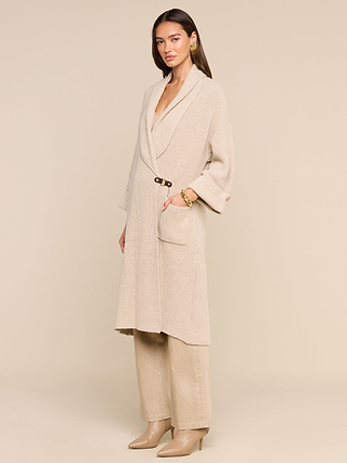 A woman stands in a beige room wearing the Orielle Cardigan—a long, light beige knit with a belt—paired with matching pants and heeled boots. Perfect for fall sweater weather, her cozy wool cardigan enhances the tranquil ambiance as she slips one hand into her pocket and gazes softly to the side.