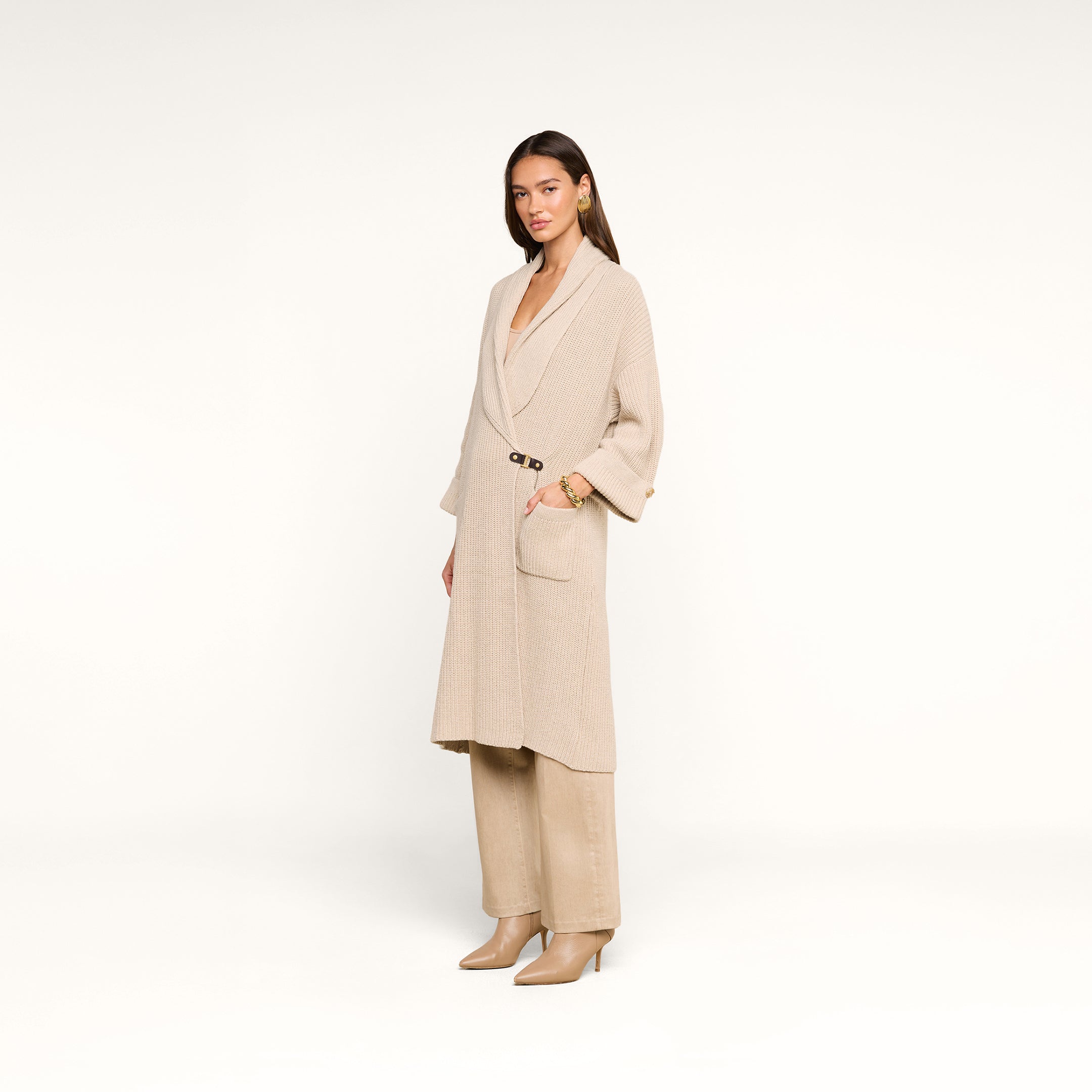 A woman poses against a white background in a beige long coat, beige pants, and beige heeled boots. She has straight brown hair and minimal accessories, with the Orielle Cardigan subtly layered under her coat for added warmth.
