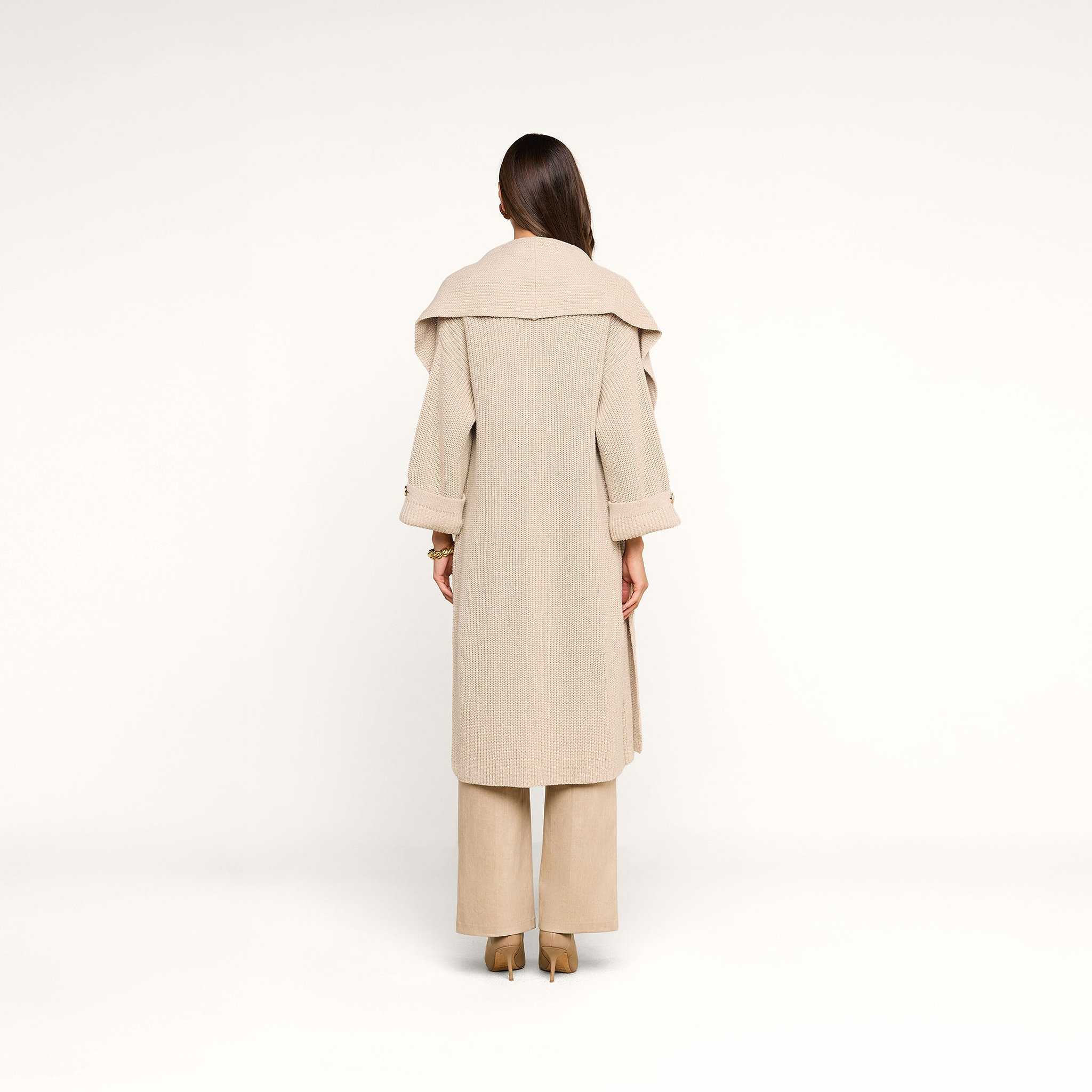 A woman with long brown hair stands facing away, wearing the Orielle Cardigan—beige with a wide collar—paired with beige wide-leg pants and brown heels against a plain white background.