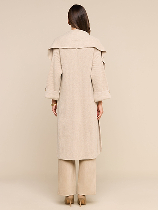 A woman stands facing away, wearing the Orielle Cardigan—a long, beige textured knit reminiscent of a cozy fall sweater with rolled sleeves—paired with wide-legged pants and high-heeled shoes. The background is a solid, light beige color.