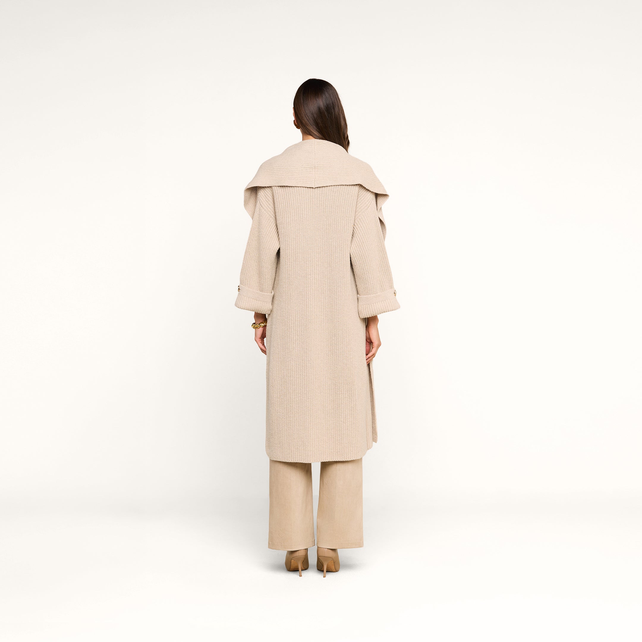 A woman with long brown hair stands facing away, wearing the Orielle Cardigan—beige with a wide collar—paired with beige wide-leg pants and brown heels against a plain white background.