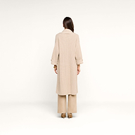 A woman stands facing away, wearing a long beige coat layered over the Orielle Cardigan, wide-leg beige pants, and beige heels, against a plain white background.