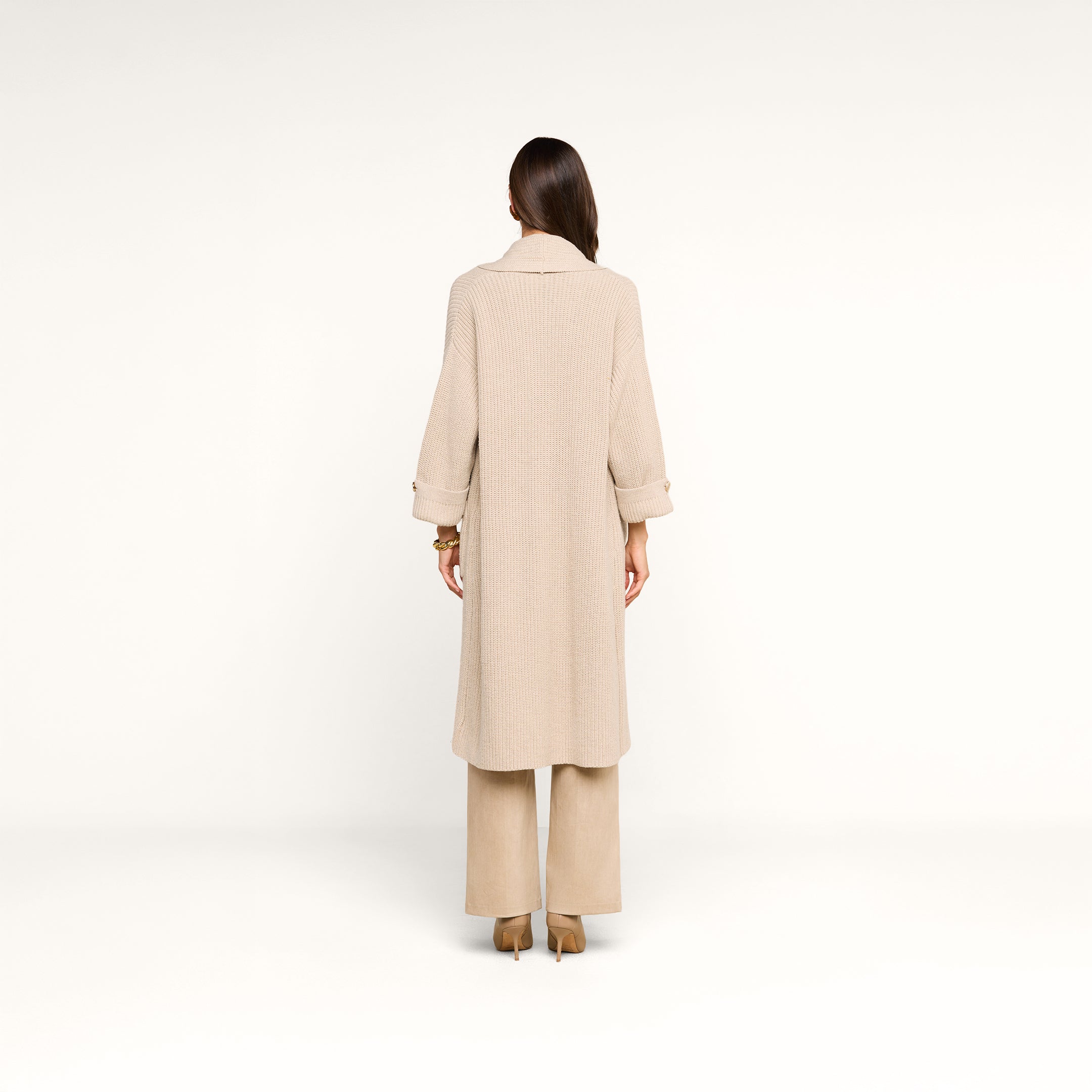 A woman stands facing away, wearing a long beige coat layered over the Orielle Cardigan, wide-leg beige pants, and beige heels, against a plain white background.