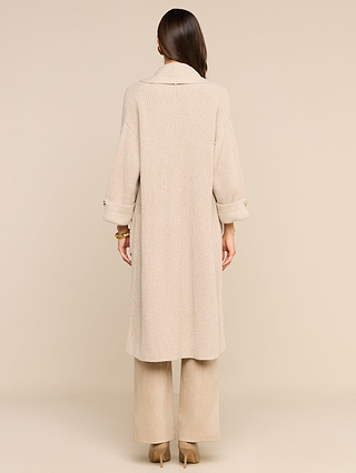 A woman stands facing away against a plain beige background, wearing the Orielle Cardigan—a long, light beige ribbed wool piece—layered over wide-leg beige pants and tan heels. Her hair is straight and she wears bracelets on her left wrist.