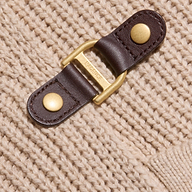 Close-up of a gold and brown leather buckle fastener attached to the Orielle Cardigan, a beige wool knit. The metal part is engraved with the word TRENERY.
