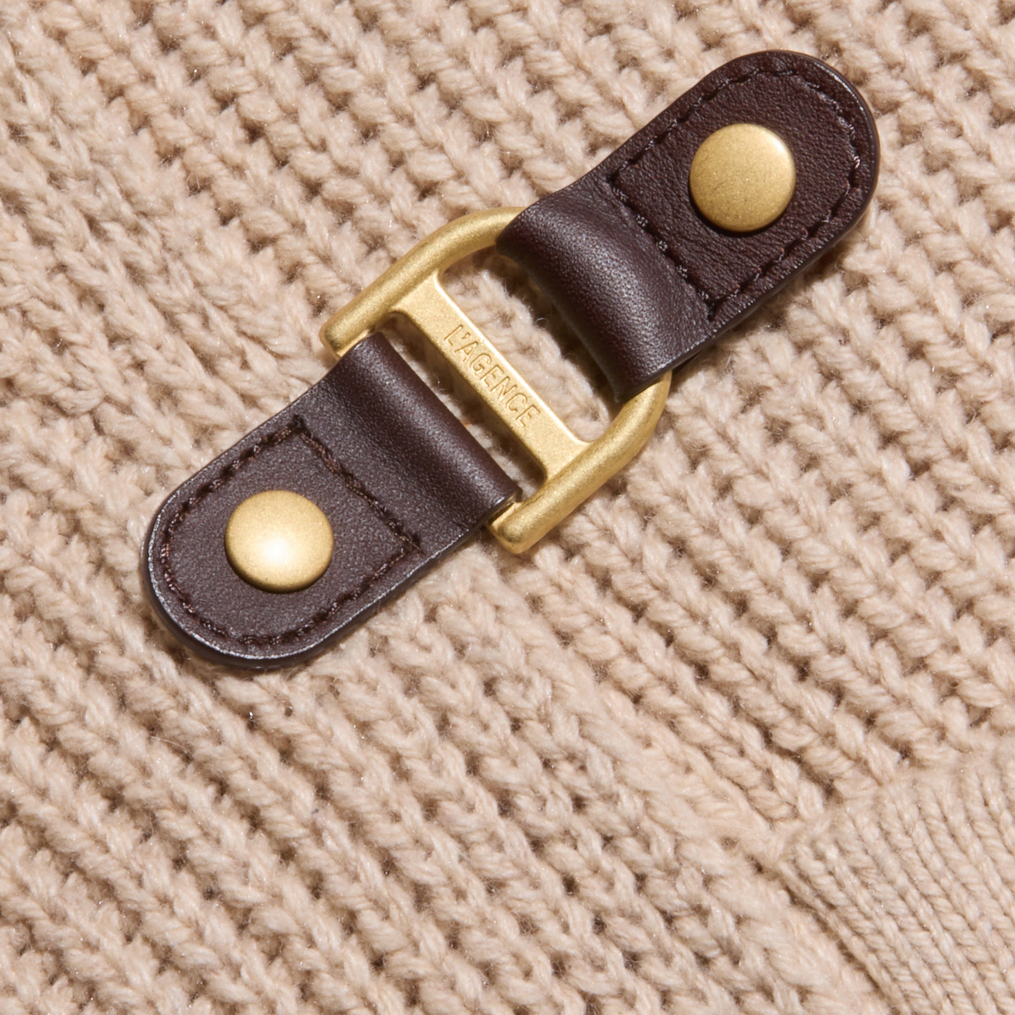 Close-up of a gold and brown leather buckle fastener attached to the Orielle Cardigan, a beige wool knit. The metal part is engraved with the word TRENERY.