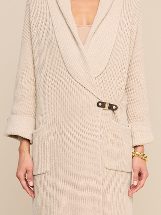 A person is wearing the Orielle Cardigan in beige, featuring large lapels, front pockets, a single buckle closure, and rolled sleeves. They have accessorized with a gold chain bracelet on their right wrist.