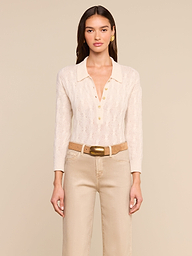 A woman with long brown hair stands against a plain background, wearing the Airy Cable Knit Sweater—a cream-colored vintage-inspired piece featuring gold buttons and a collared neckline—paired with beige pants and a tan belt with a large gold buckle. She is looking straight ahead.