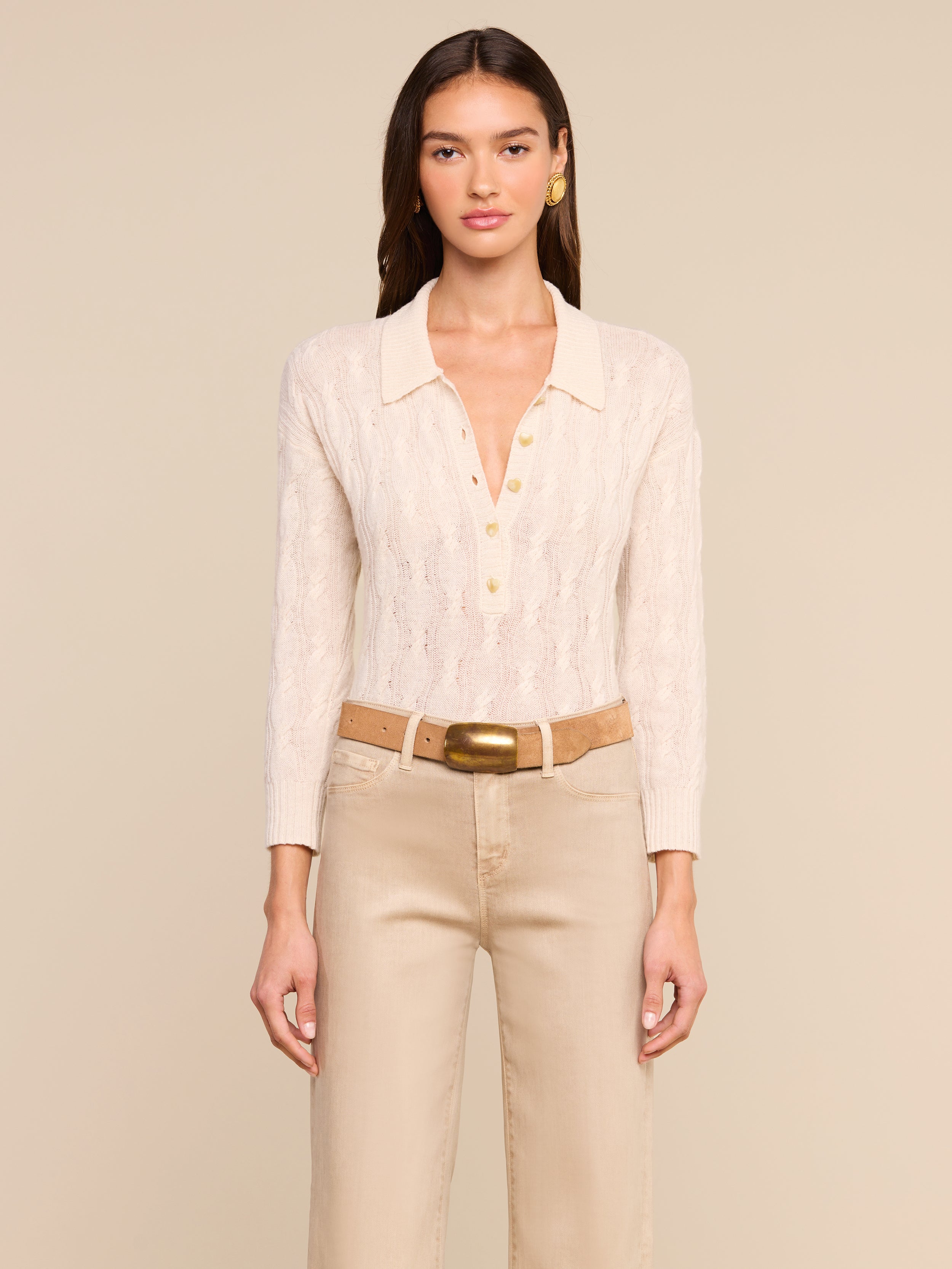 A woman with long brown hair stands against a plain background, wearing the Airy Cable Knit Sweater—a cream-colored vintage-inspired piece featuring gold buttons and a collared neckline—paired with beige pants and a tan belt with a large gold buckle. She is looking straight ahead.