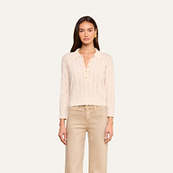 A woman stands facing forward against a plain light background, wearing the Airy Cable Knit Sweater in a cream color with heart-shaped buttons, paired with beige pants.