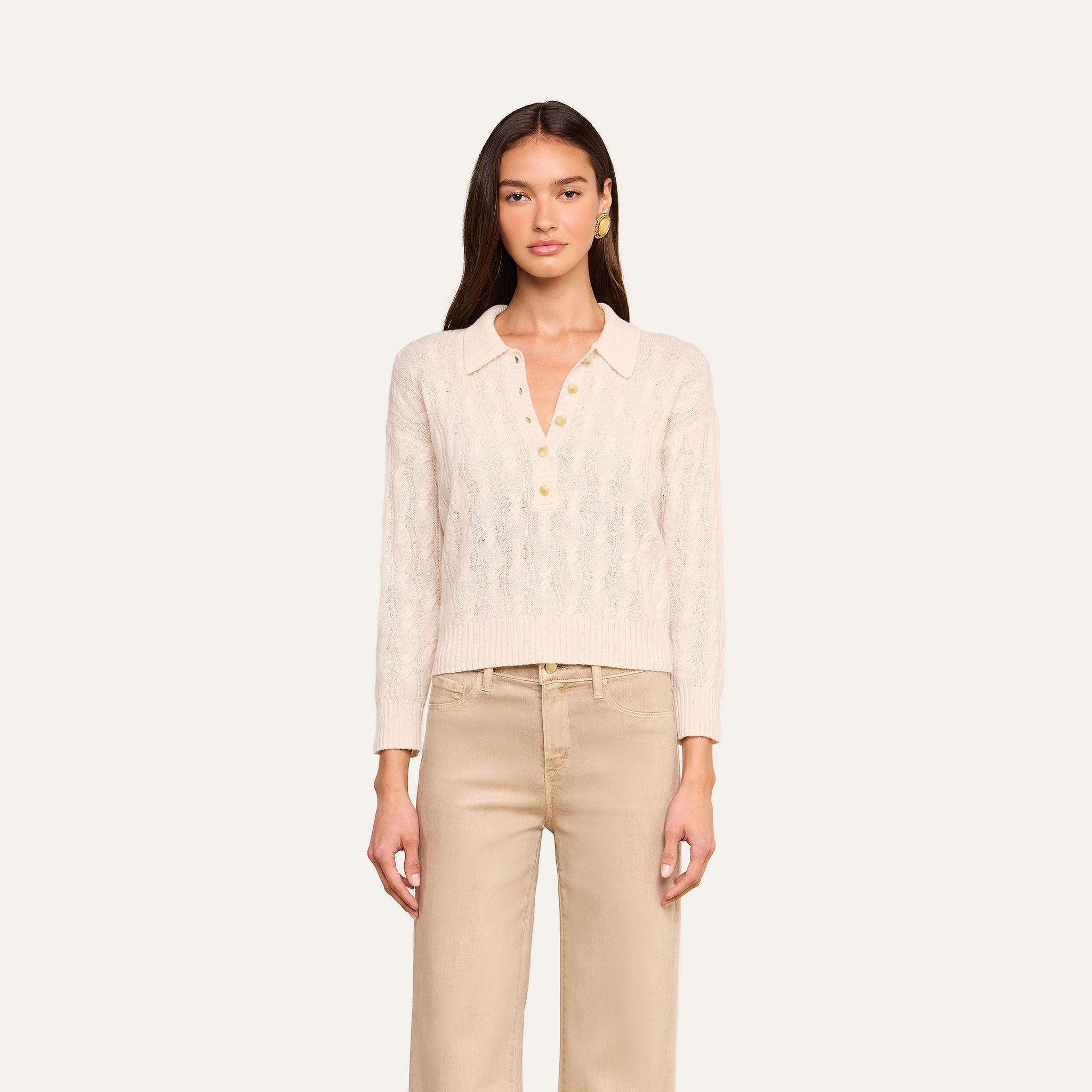 A woman stands facing forward against a plain light background, wearing the Airy Cable Knit Sweater in a cream color with heart-shaped buttons, paired with beige pants.
