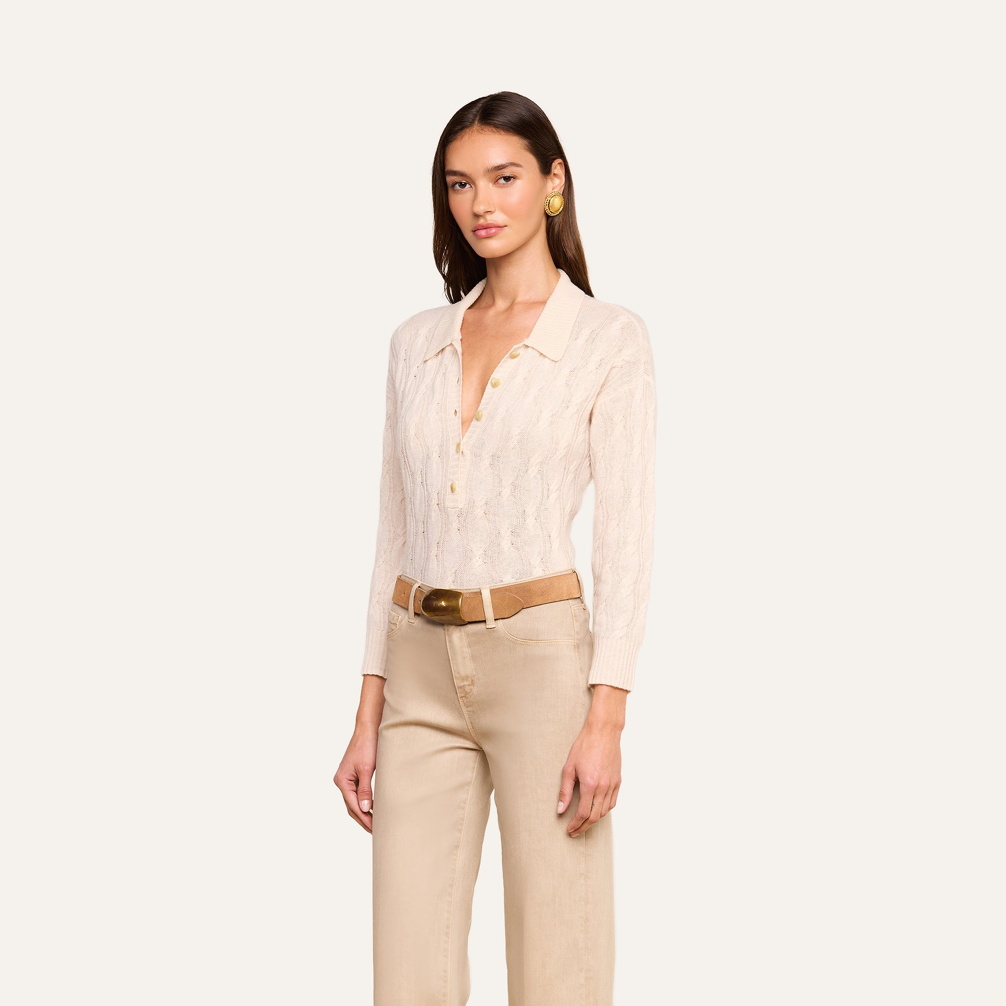 A woman stands against a plain background wearing the Airy Cable Knit Sweater, a vintage beige collared style with heart buttons. She pairs it with beige pants, a tan belt, and gold earrings. Her straight brown hair frames her neutral expression.