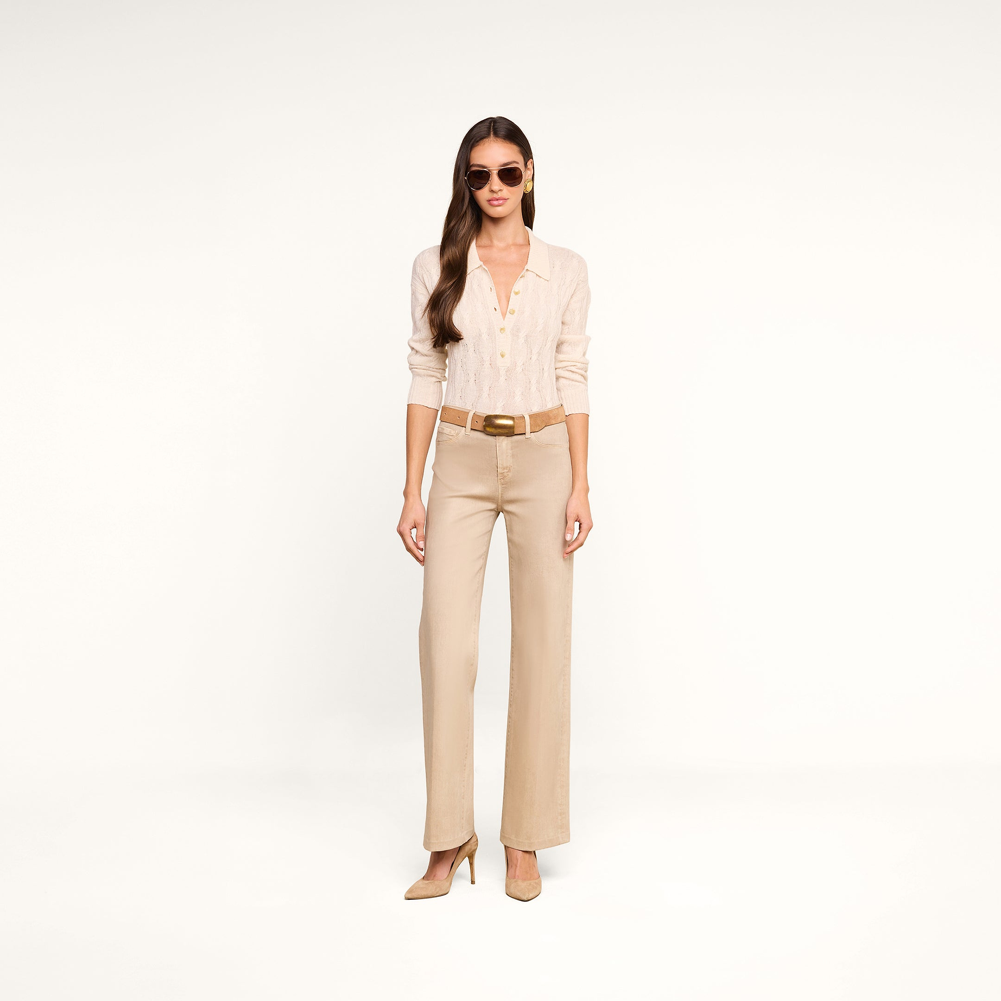 A woman wearing sunglasses and the Airy Cable Knit Sweater—a beige long-sleeve cable knit top with heart buttons—paired with tan wide-leg belted pants and tan heels stands against a plain white background.
