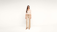A woman wearing sunglasses, a light beige blouse, wide-leg tan pants, and tan heels stands against a plain white background.