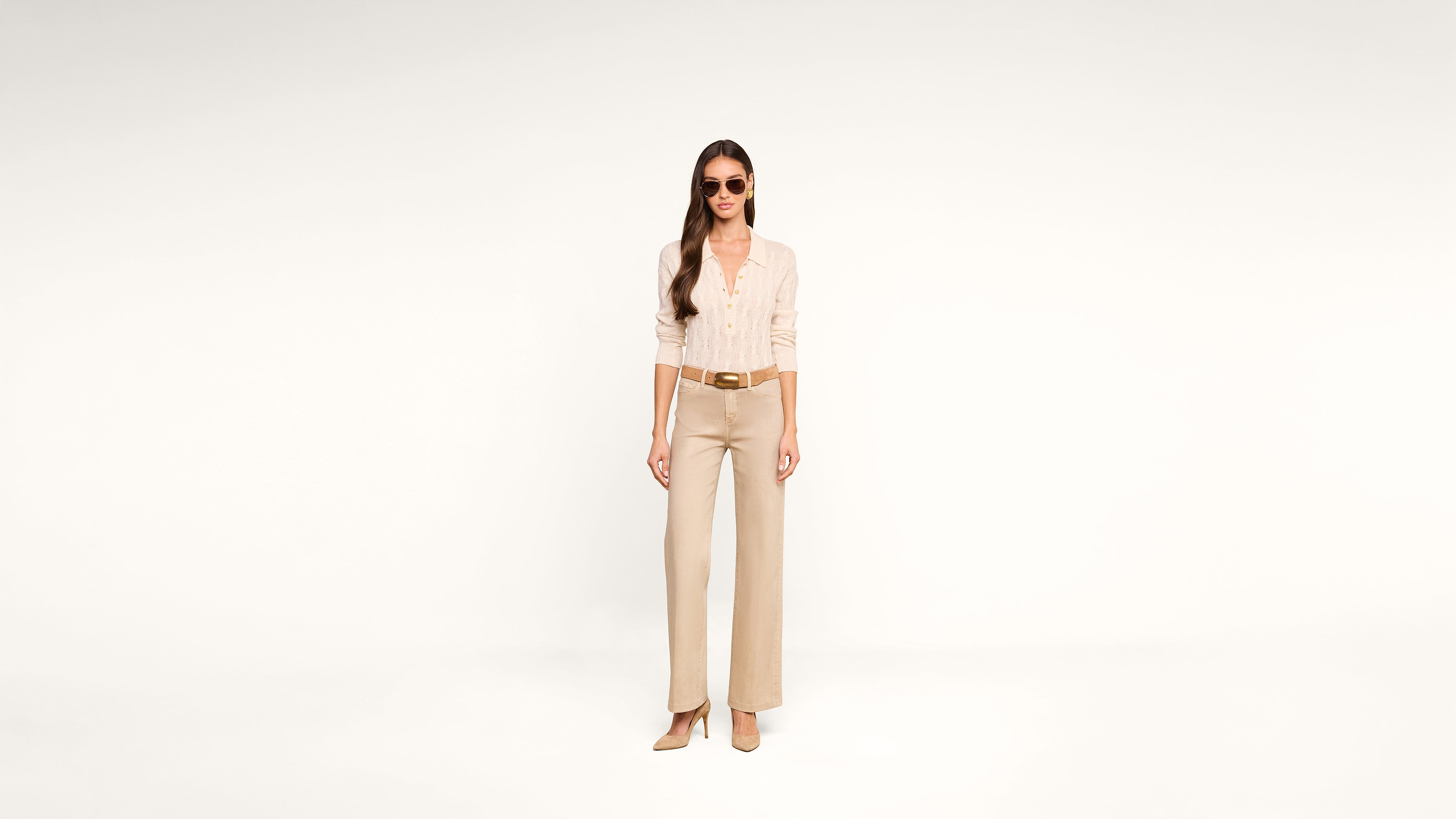 A woman wearing sunglasses, a light beige blouse, wide-leg tan pants, and tan heels stands against a plain white background.