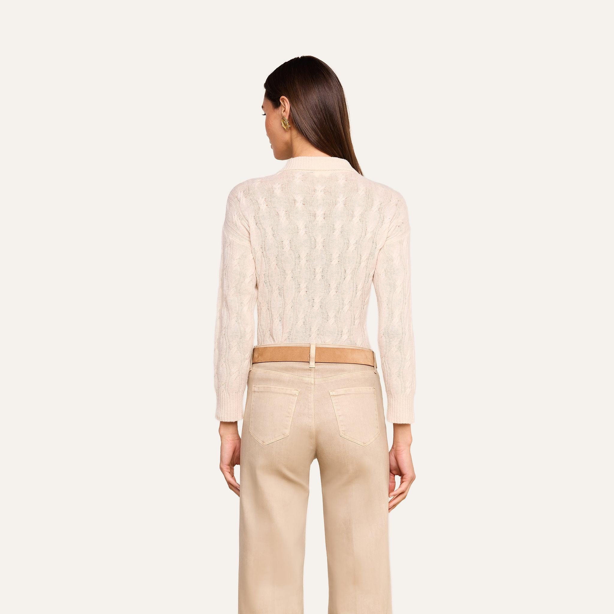 A woman stands with her back to the camera, wearing the Airy Cable Knit Sweater in a light hue and beige pants against a plain light background. Her straight brown hair is down, and her hands rest relaxed at her sides.