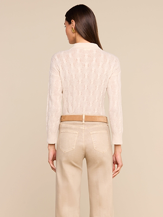 A woman stands with her back to the camera, wearing the Airy Cable Knit Sweater in a cream shade with a high collared neckline, paired with light beige pants against a plain beige background.