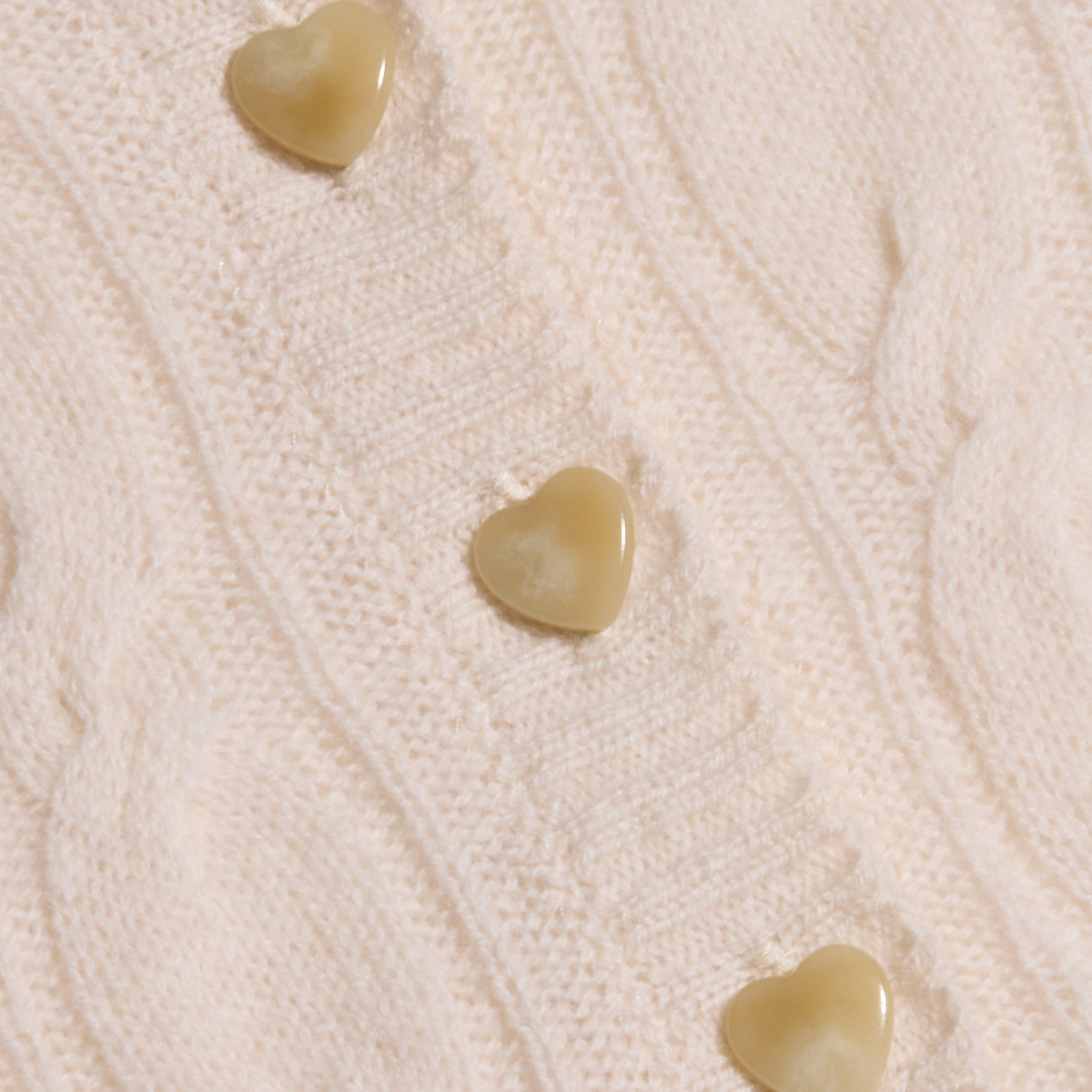 A close-up of the Airy Cable Knit Sweater in a creamy hue, showcasing three light brown, heart-shaped buttons sewn along its ribbed detail.