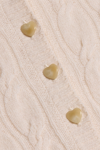 A close-up of the Airy Cable Knit Sweater in a cream shade, featuring three light yellow heart-shaped buttons arranged vertically.