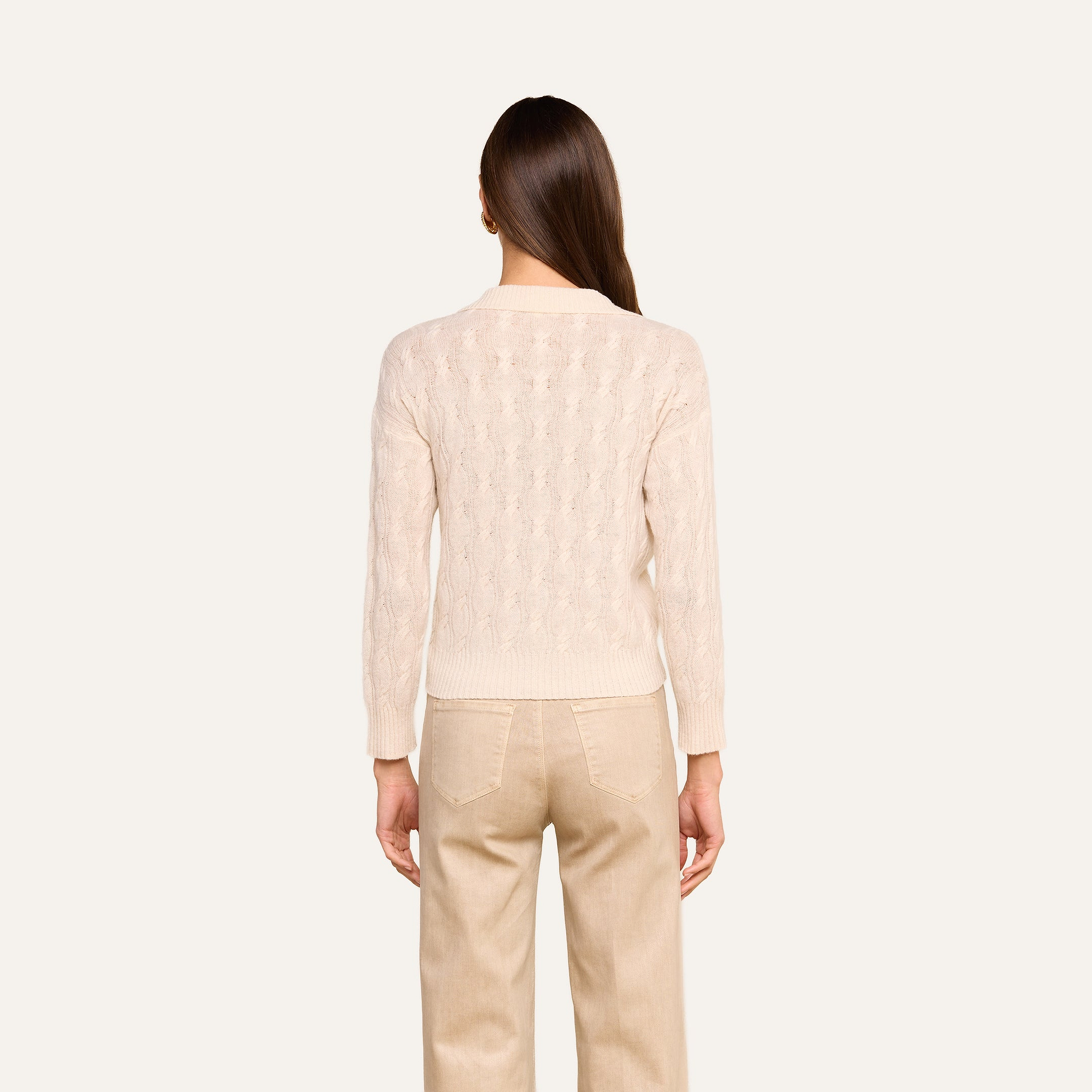 A person with long brown hair stands facing away, wearing the Airy Cable Knit Sweater in a cream shade and beige pants against a plain light background.