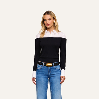 A woman wearing the Mathilde Poplin Collar Pullover, blue jeans, a black belt with a gold buckle, and gold earrings stands against a beige background, looking forward with a neutral expression.