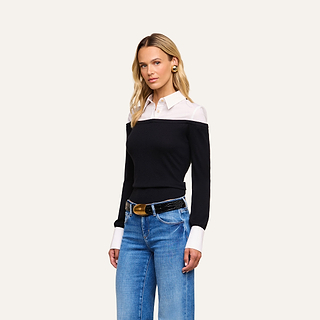 A woman stands against a beige background wearing the Mathilde Poplin Collar Pullover—a black and white long-sleeve shirt with a white collar and cuffs—paired with blue jeans, a black belt featuring a gold buckle, and gold earrings. She has long, wavy blonde hair.