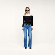 A woman stands facing forward against a plain, light background, wearing sunglasses, blue jeans, black shoes, and the Mathilde Poplin Collar Pullover in black and white.