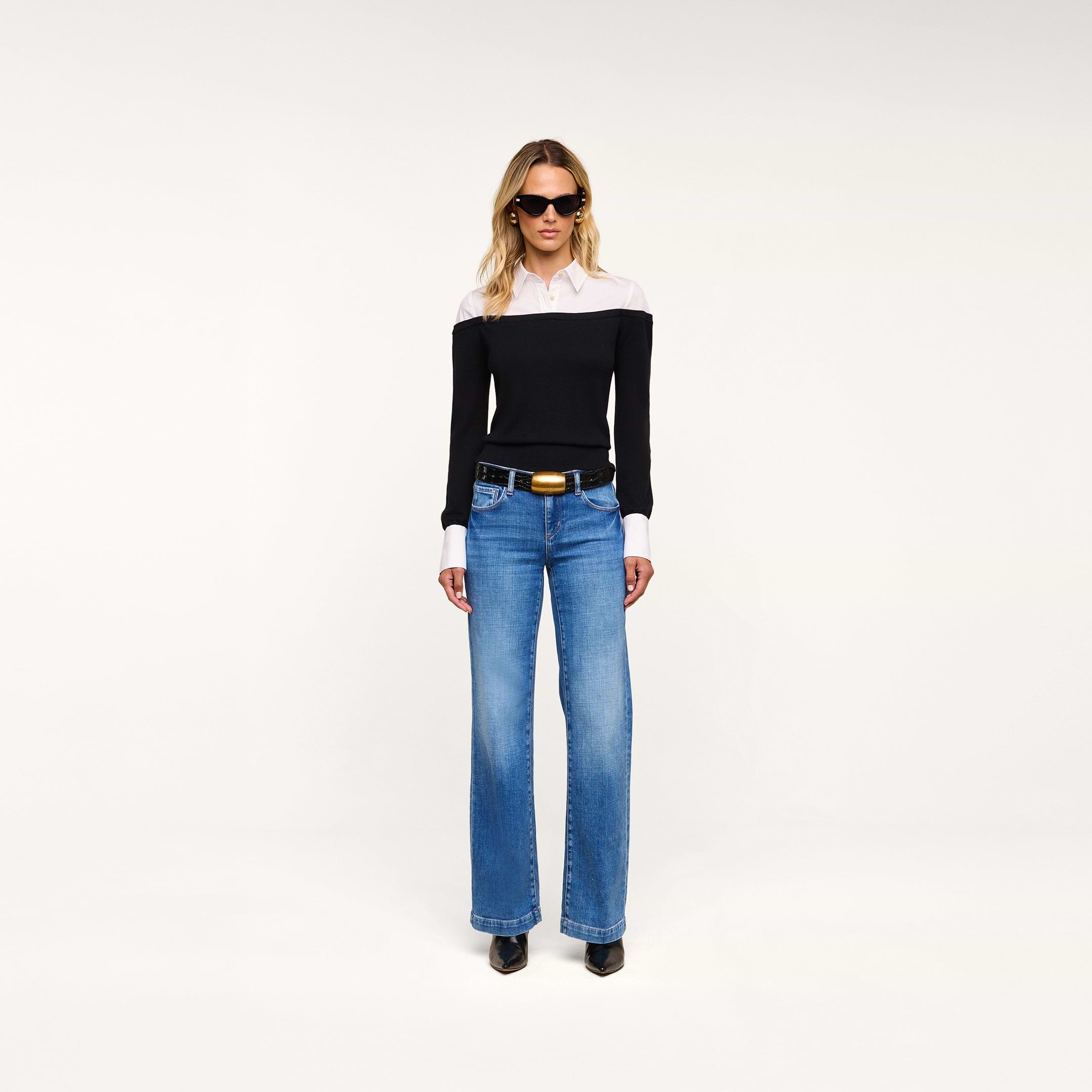 A woman stands facing forward against a plain, light background, wearing sunglasses, blue jeans, black shoes, and the Mathilde Poplin Collar Pullover in black and white.