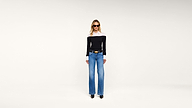 A woman wearing a black-and-white top, blue wide-leg jeans, black shoes, and sunglasses stands against a plain white background.