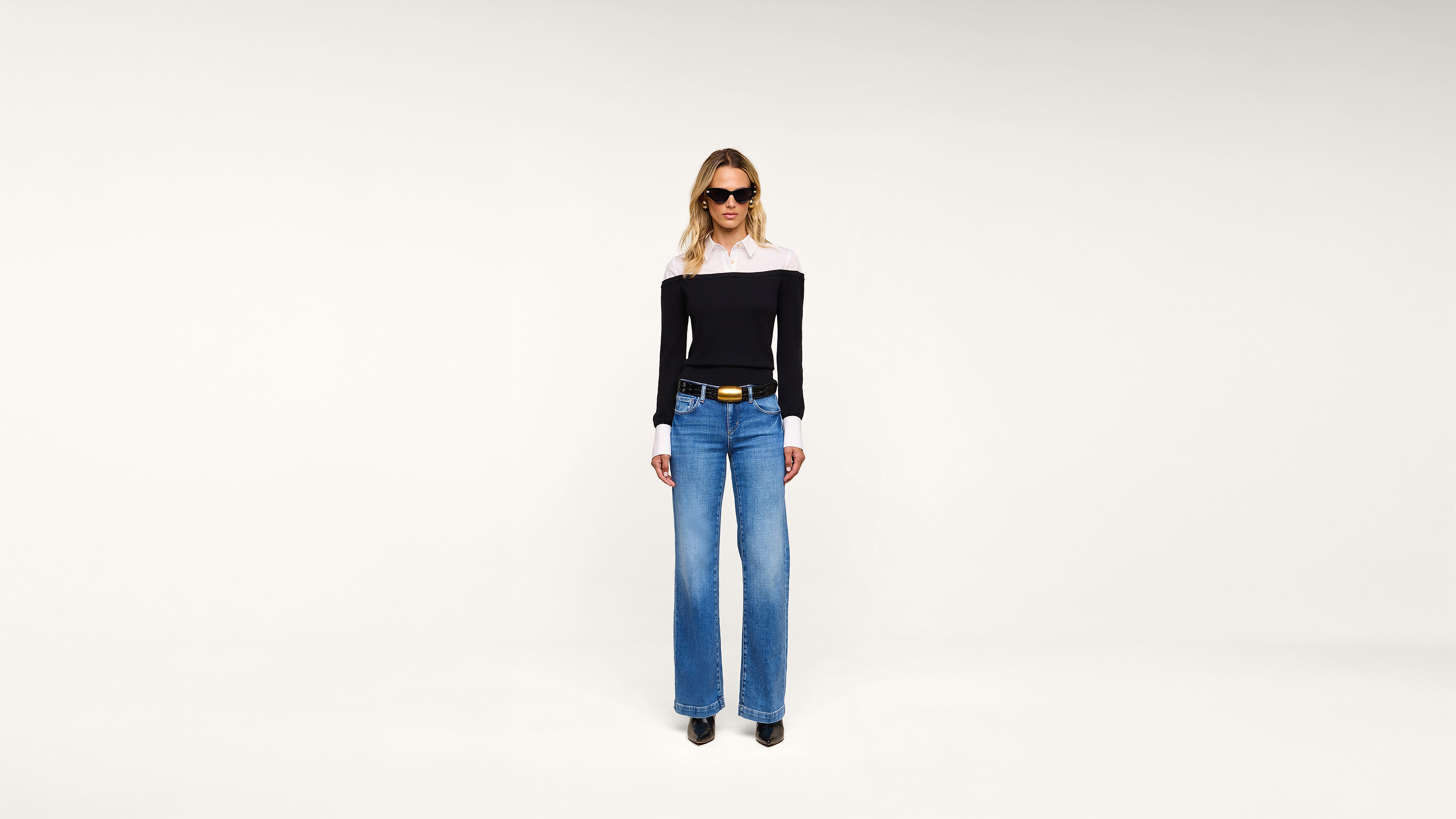 A woman wearing a black-and-white top, blue wide-leg jeans, black shoes, and sunglasses stands against a plain white background.