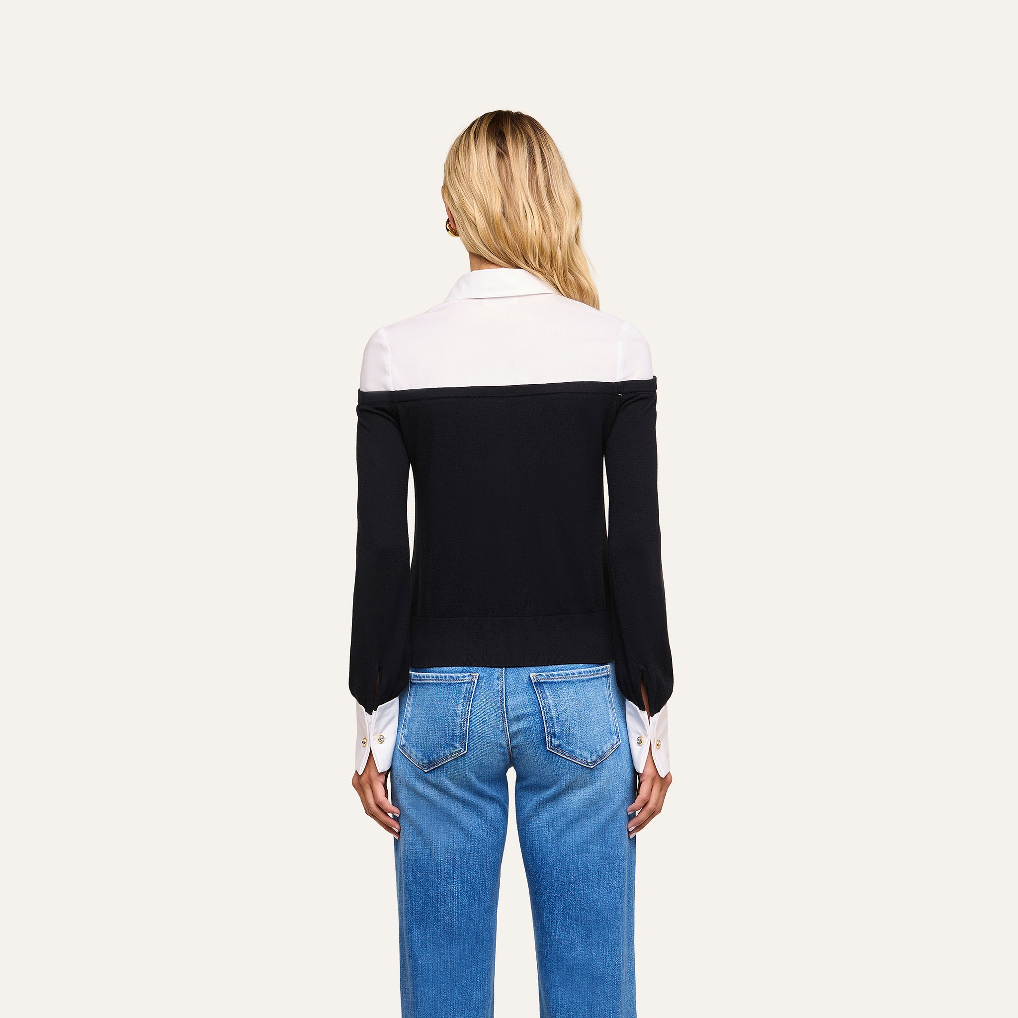 A woman stands facing away, wearing the Mathilde Poplin Collar Pullover—featuring a black and white design with white collar and cuffs—paired with blue jeans, set against a plain light background.