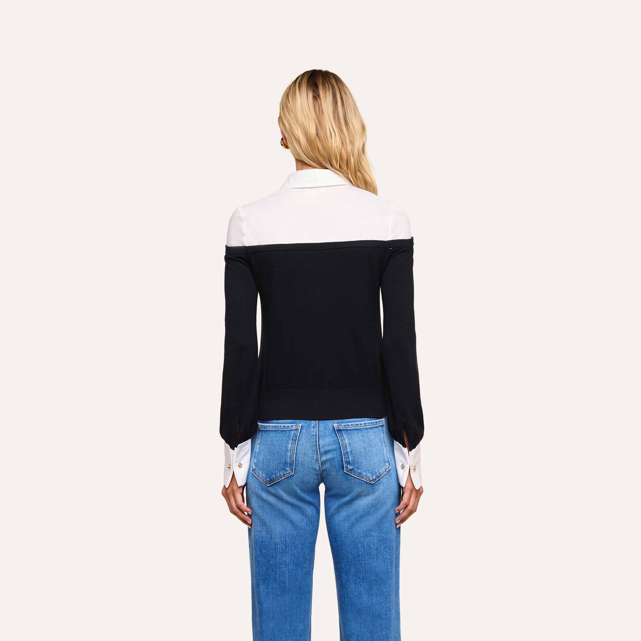 A woman stands facing away, wearing the Mathilde Poplin Collar Pullover—featuring a black and white design with white collar and cuffs—paired with blue jeans, set against a plain light background.