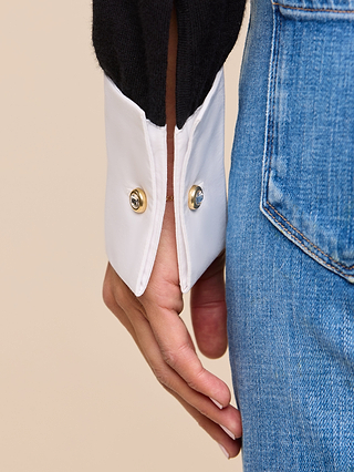 A close-up of a person’s hand resting near the pocket of their denim jeans as they wear the Mathilde Poplin Collar Pullover, featuring off-the-shoulder styling and white cuffs secured with two gold buttons.