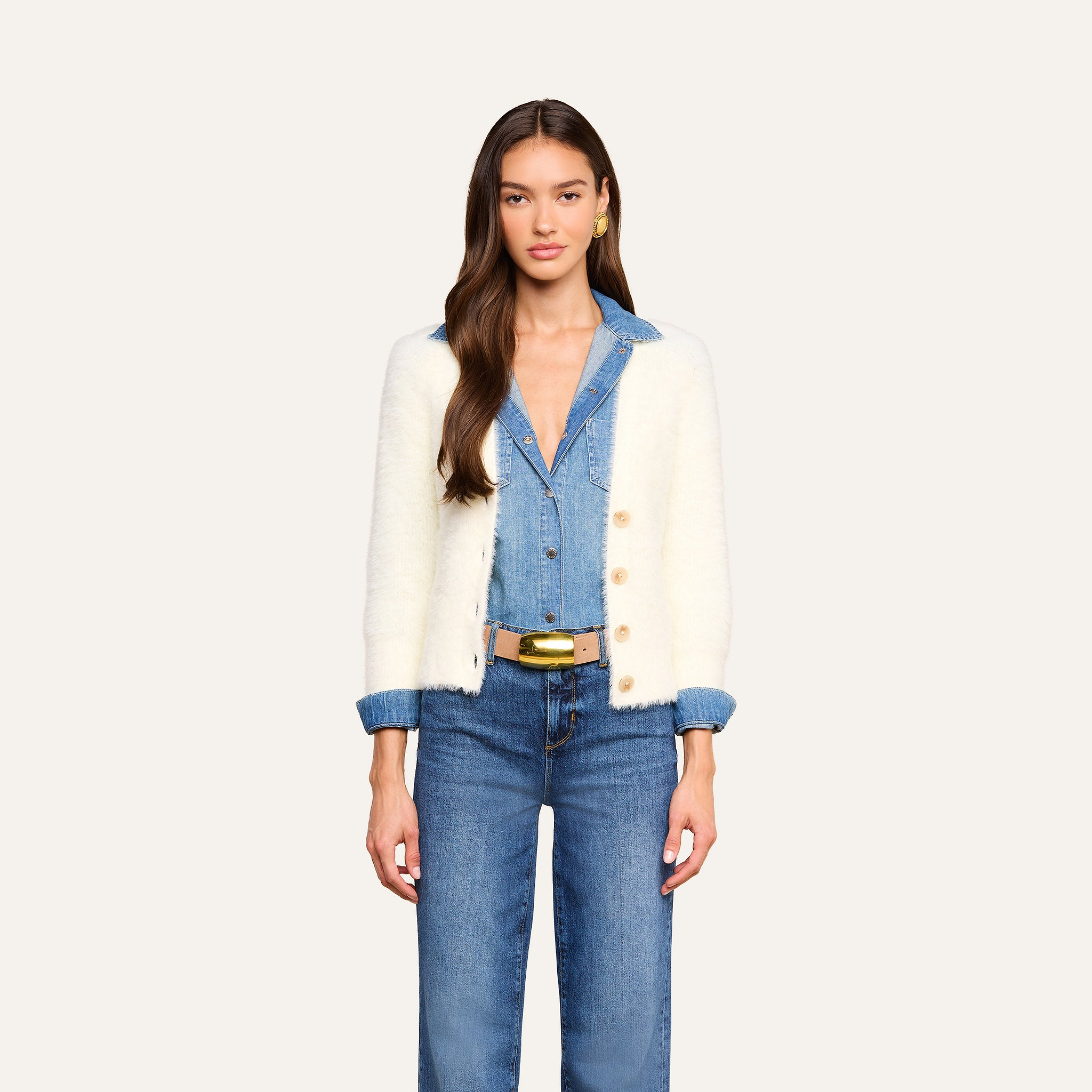 Una donna è in piedi, rivolta verso lobiettivo, e indossa il cardigan Agathe sopra una camicia di jeans blu e dei jeans. Ha lunghi capelli castani e orecchini doro, in risalto su uno sfondo beige a tinta unita.