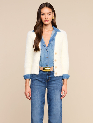 A woman stands facing forward, wearing the Agathe Cardigan Top over a blue denim shirt and jeans. She has long brown hair and gold earrings, set against a plain beige background.