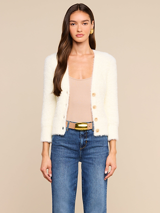 A woman wearing the Agathe Cardigan Top over a beige v-neck top, paired with blue jeans, a wide belt with a gold buckle, and gold earrings stands in front of a plain beige background.
