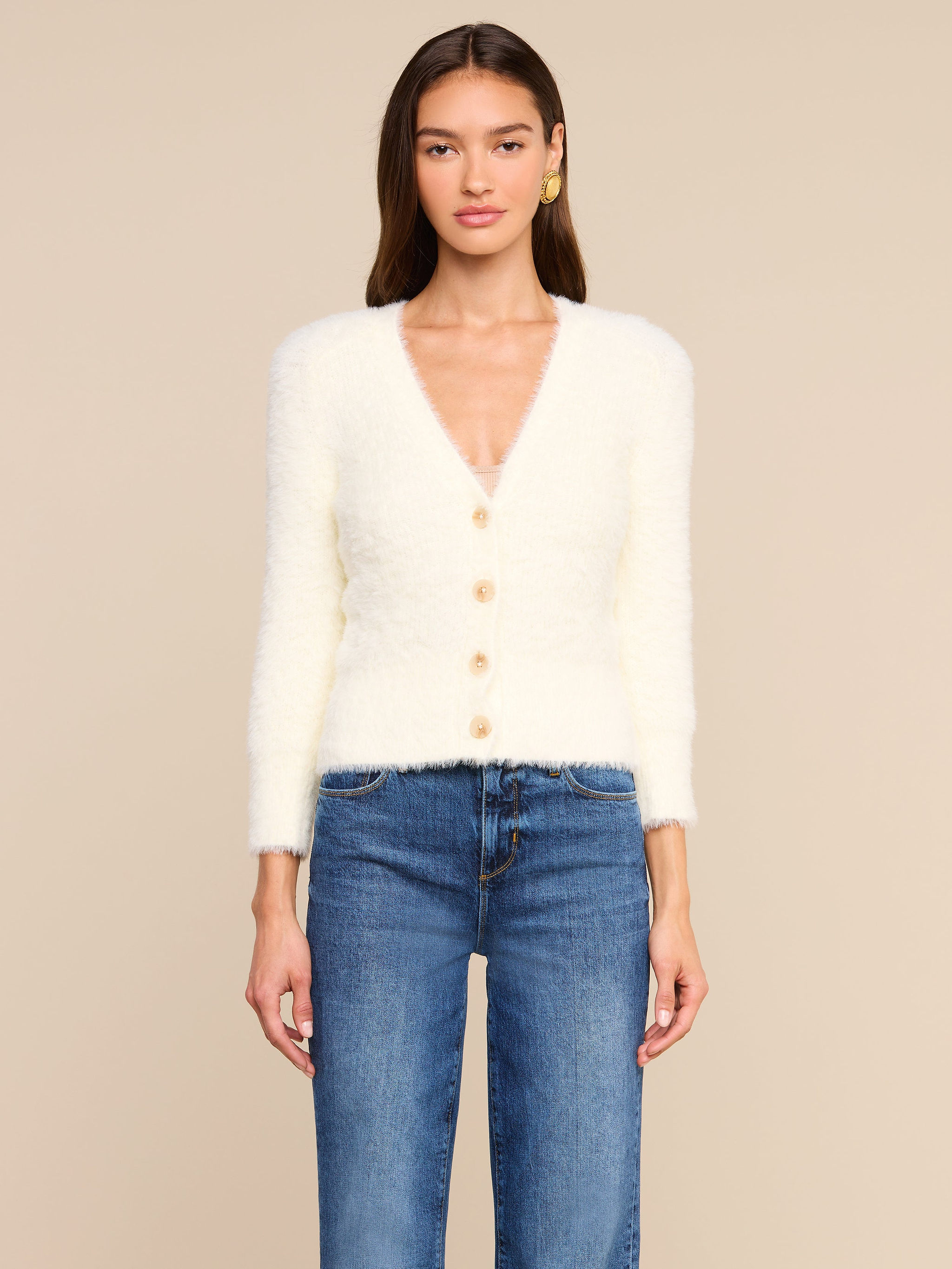 Una donna è in piedi, rivolta verso lobiettivo, e indossa lAgathe Cardigan Top, un cardigan morbido con scollo a V e bottoni, abbinato a un paio di jeans blu. Ha lunghi capelli castani e orecchini doro. Lo sfondo è beige.
