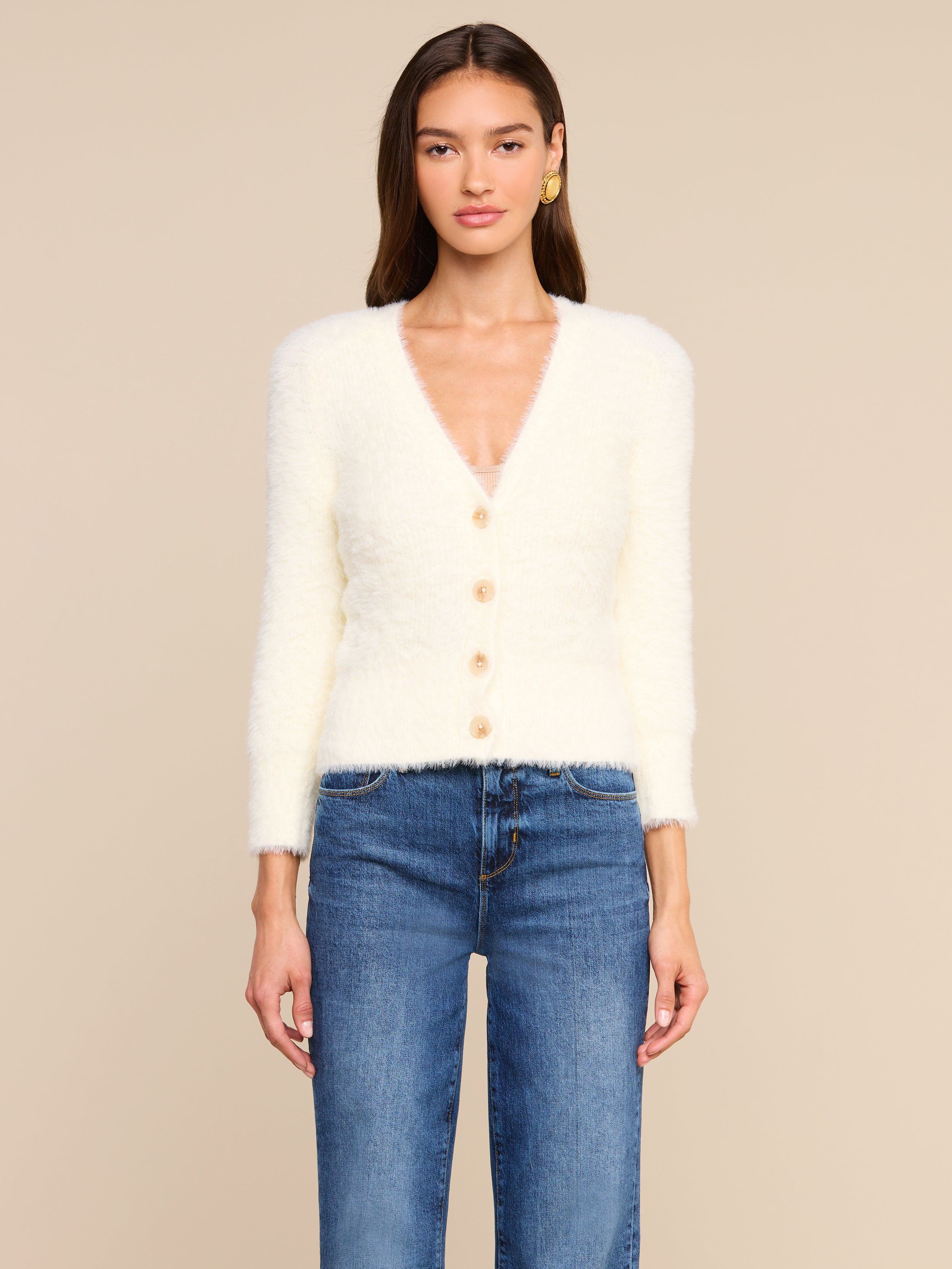 Una donna è in piedi, rivolta verso lobiettivo, e indossa lAgathe Cardigan Top, un cardigan morbido con scollo a V e bottoni, abbinato a un paio di jeans blu. Ha lunghi capelli castani e orecchini doro. Lo sfondo è beige.