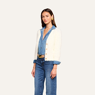 Una donna è in piedi su uno sfondo beige a tinta unita e indossa il cardigan Agathe sopra una camicia di jeans e dei jeans, con una cintura e grandi orecchini doro.