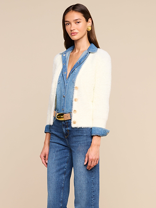 Una donna è in piedi su uno sfondo beige a tinta unita e indossa il cardigan Agathe sopra una camicia di jeans e dei jeans, con una cintura e grandi orecchini doro.