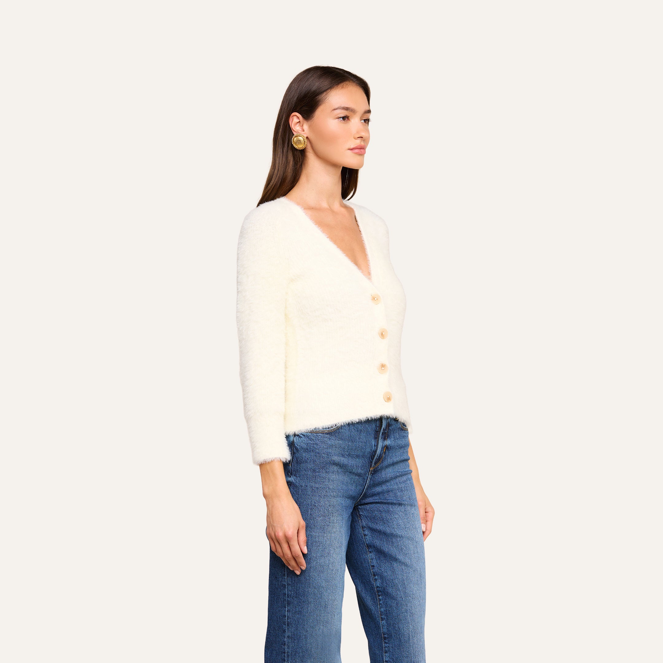 Una donna che indossa il cardigan Agathe — un cardigan corto, bianco e morbido, con maniche lunghe — abbinato a jeans blu, è in piedi su uno sfondo beige, rivolta verso sinistra con un’espressione neutra.