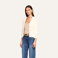 Una donna che indossa l’Agathe Cardigan Top — un cardigan corto bianco e soffice — abbinato a un top beige con scollo a V, jeans blu e orecchini dorati, è in posa davanti a uno sfondo beige a tinta unita.