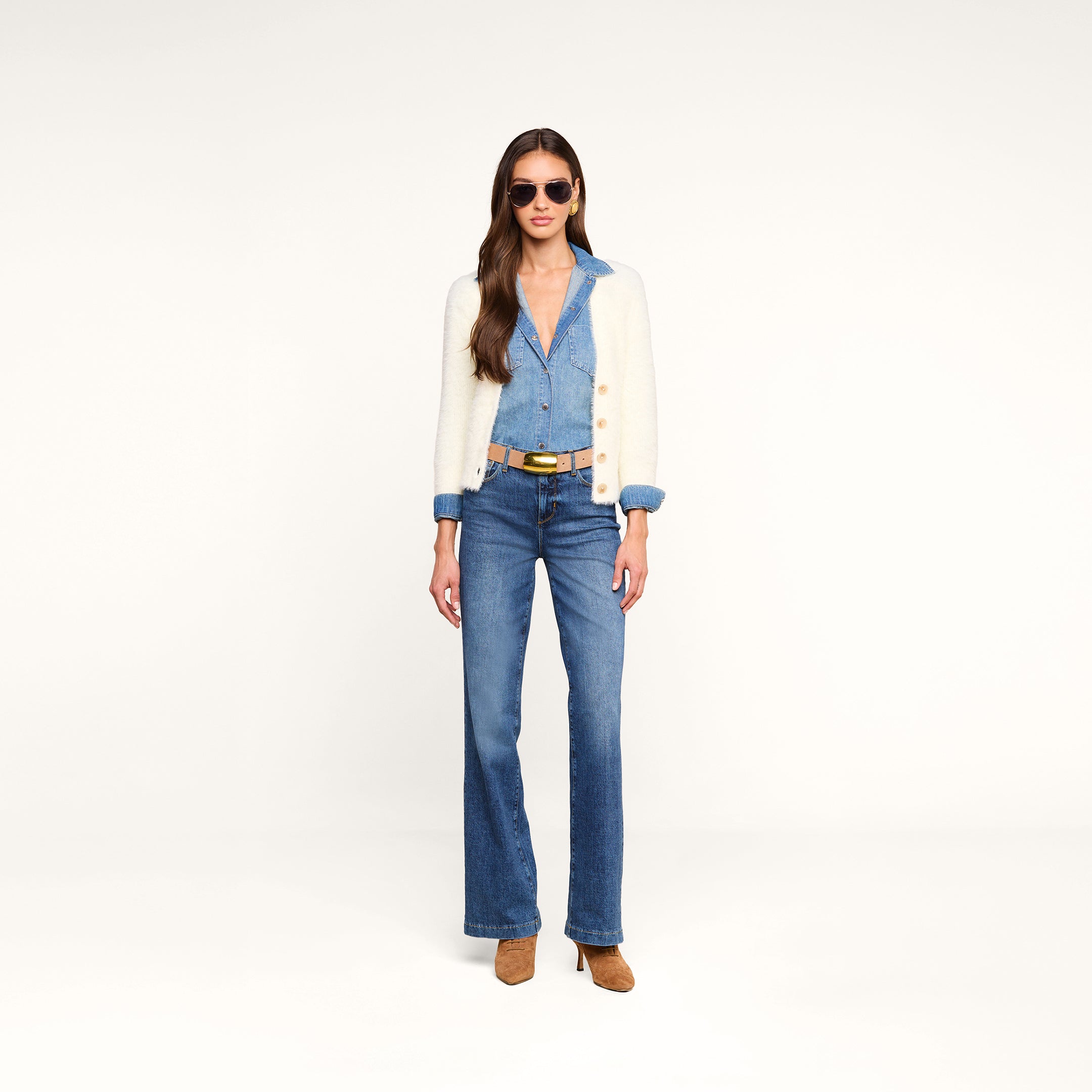 Una donna con indosso occhiali da sole, il cardigan “Agathe”, una tuta in denim con una cintura beige e scarpe con tacco color cuoio è in piedi davanti a uno sfondo beige a tinta unita, con lo sguardo rivolto in avanti e le mani lungo i fianchi.