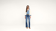 Woman wearing sunglasses, a light beige cardigan, a blue button-up shirt, blue high-waisted jeans, a brown belt, and brown shoes stands against a plain white background.